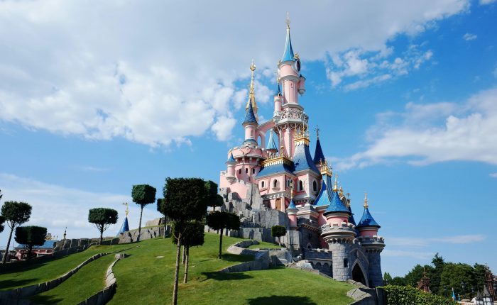 Pink castle with blue roofs and gold spires perched on a grassy hill under a bright blue sky.