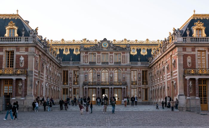Front view of a grand palace with gilded sculptures, blue slate roof, and decorative stone details, with visitors on the cobblestone foreground.