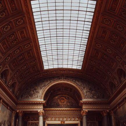 Interior of a grand historic gallery with a glass skylight, ornate gilded ceiling, marble columns, and a large central painting; visitors at the front.